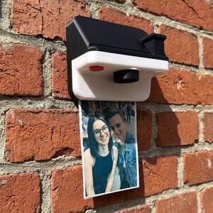 May include: A black and white 3D-printed camera-shaped photo holder fixed to a red brick wall. A printed photo of a couple is displayed below the camera. The camera features a red button and a lens.