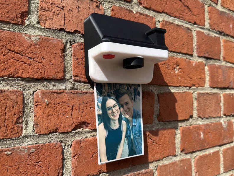 May include: A black and white 3D-printed camera-shaped photo holder fixed to a red brick wall. A printed photo of a couple is displayed below the camera. The camera features a red button and a lens.