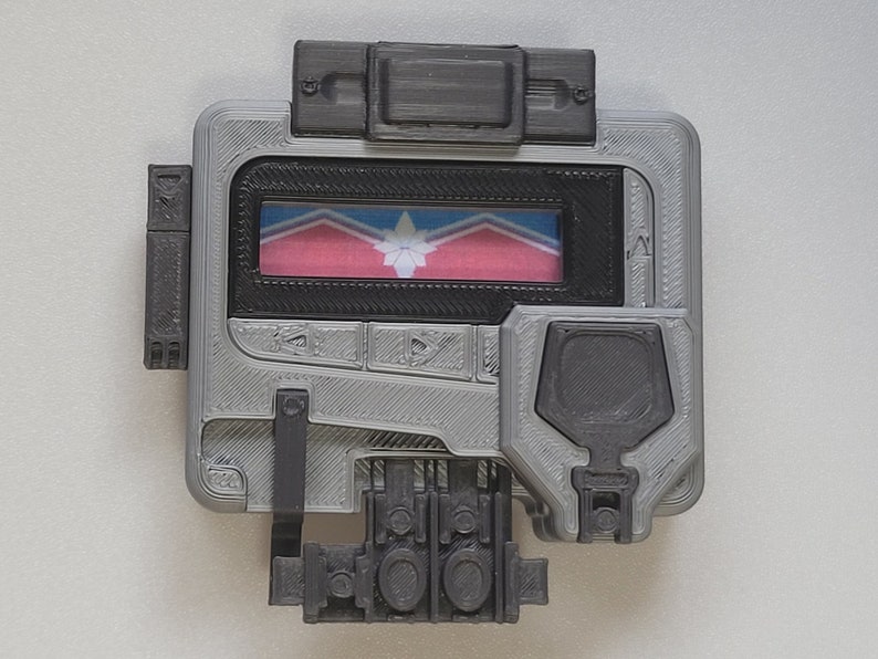 May include: A gray and black handheld device with a screen displaying a pink, blue, and white flag. The device has several buttons and a small speaker.