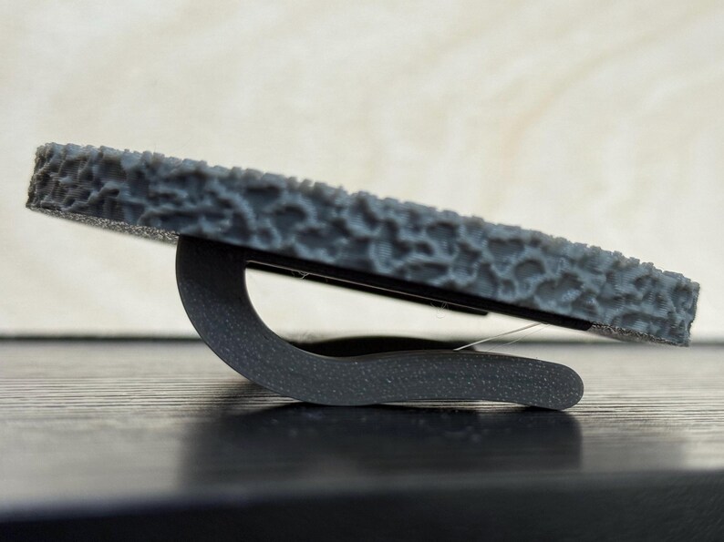 Puede incluir: Un soporte para tel&eacute;fono gris oscuro texturizado con una base curva. El soporte est&aacute; dise&ntilde;ado para sostener un tel&eacute;fono o tableta, proporcionando una experiencia de visualizaci&oacute;n estable e inclinada. El soporte est&aacute; hecho de un material duradero.