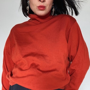 Könnte beinhalten: Ein rostfarbener Rollkragenpullover mit langen Ärmeln. Der Pullover wird mit dunkelblauen Hosen getragen. Das Model trägt silberne Ohrringe und hat dunkles Haar.