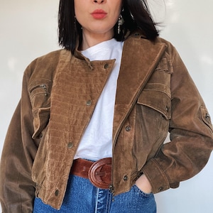 Könnte beinhalten: Braune Wildlederjacke mit Reißverschluss, mehreren Taschen und Knöpfen. Die Jacke wird mit einem braunen Ledergürtel und einer blauen Jeans kombiniert. Die Jacke hat einen klassischen, Vintage-Stil.