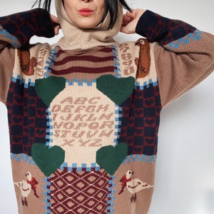 Peut inclure: Un pull à col montant et à capuche, de couleur marron, vert, bleu et blanc, avec un motif patchwork composé de différentes formes géométriques et un motif d'oiseau.
