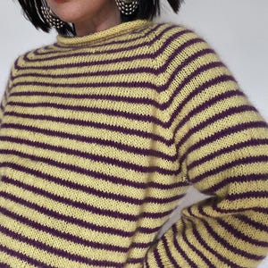 Puede incluir: Un su&eacute;ter a rayas amarillas y moradas con cuello alto. El su&eacute;ter presenta rayas horizontales en un patr&oacute;n repetitivo. La prenda de punto parece suave y c&oacute;moda, adecuada para climas m&aacute;s fr&iacute;os. Las mangas son largas y el ajuste es holgado.
