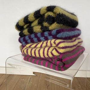 Puede incluir: Una pila de cuatro gorros de punto doblados con rayas horizontales en varios colores. Los gorros est&aacute;n apilados sobre una mesa auxiliar de acr&iacute;lico transparente. Los colores incluyen verde oliva y negro, morado y azul, amarillo y morado, y marr&oacute;n y rosa.