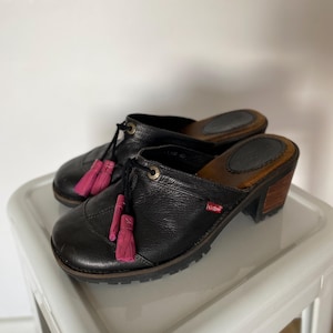 Könnte beinhalten: Schwarze Lederclogs mit rosa Quasten. Die Clogs haben einen kleinen Absatz und eine runde Spitze. Der Absatz ist ca. 5 cm hoch. Kleines rotes Logo an der Seite.