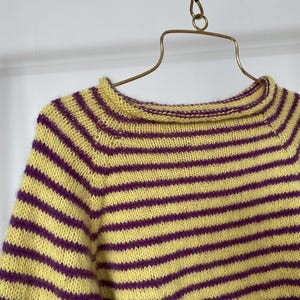 Puede incluir: Un su&eacute;ter a rayas amarillas y moradas se muestra en una percha dorada. El su&eacute;ter presenta rayas horizontales en un patr&oacute;n repetitivo. Se ven el cuello y las mangas, mostrando la construcci&oacute;n de punto y la combinaci&oacute;n de colores.