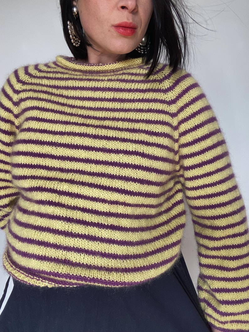 Puede incluir: Un jersey a rayas amarillas y moradas. El jersey presenta rayas horizontales en amarillo claro y morado oscuro. El cuello y los pu&ntilde;os son de color morado liso. El jersey es de punto.