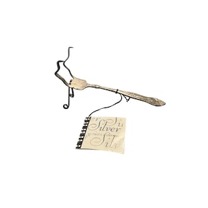 May include: A silver-toned metal fork bent into a decorative shape, supported by a black wire stand. A black string attaches a rectangular tag with the text Silver by Two's Company to the handle of the fork, which features an embossed pattern.