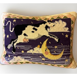 May include: A rectangular needlepoint pillow featuring a black and white cow jumping over a yellow crescent moon against a dark purple night sky with white clouds and yellow stars. The border has a repeating green triangle pattern on a cream background.