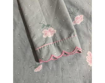 Puede incluir: Una sábana de tela gris claro con un patrón repetitivo de pequeñas flores blancas y motivos florales rosas más grandes con tallos verdes. El borde de la tela está acabado con un ribete festoneado rosa y una fina línea horizontal rosa.