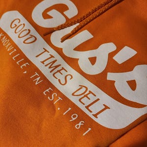 May include: Orange hoodie with white text that reads "Gus's Good Times Deli Knoxville, TN Est. 1981." The hoodie has a drawstring and a soft, comfortable appearance. The design is centered on the front of the hoodie.