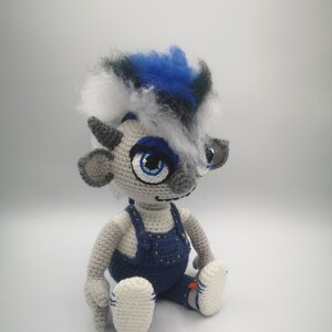 May include: A gray crocheted plush toy with blue hair, blue eyes, and a blue and white outfit. The toy is sitting with its legs crossed.