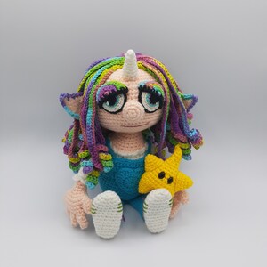 Könnte beinhalten: Eine gehäkelte Einhornpuppe mit Regenbogenhaar, die eine blaue Latzhose trägt und einen gelben Stern hält.