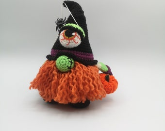 May include: A crocheted Halloween gnome with a black witch hat, a green and purple striped hat band, a large green eye, and an orange beard. The gnome is holding a small orange pumpkin.