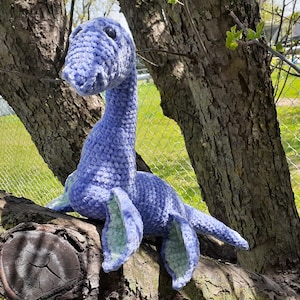 May include: A crocheted plush toy shaped like a plesiosaur, a long-necked marine reptile. The toy is light blue with green accents and is sitting on a tree branch.