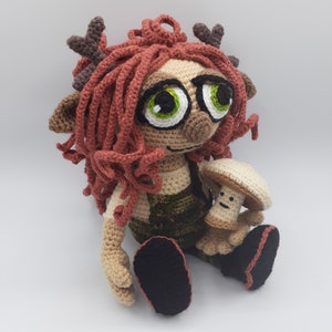 May include: A crocheted plush toy with red hair, brown antlers, and green eyes wearing a green and brown outfit and holding a white crocheted mushroom.
