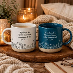 May include: Two ceramic mugs, one cream and one dark blue, each featuring a black line-art floral pattern at the base and the text: A quote you love, styled in a charming vintage typewriter font. Optional Author. Both mugs have a natural tan base.