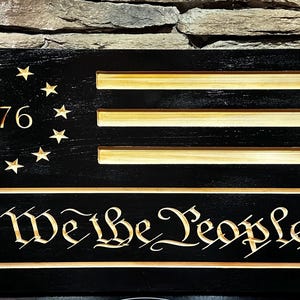 May include: A wooden sign featuring a black background with carved gold-colored stars and stripes, and the text "1776" and "We the People". The sign is designed to resemble the American flag.