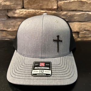May include: Gray trucker hat with a black mesh back and a curved brim. A dark cross is centered on the front. The hat has a Richardson 112 Trucker tag.
