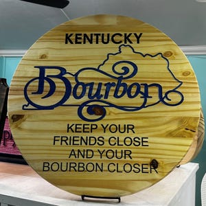 Op de afbeelding: Ronde houten plank met het woord "Bourbon" in blauw, met de omtrek van Kentucky erboven. Op het bord staat: "Houd je vrienden dichtbij en je bourbon dichterbij."