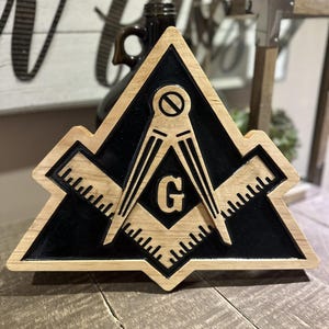 May include: Wooden Masonic symbol with a black center and light brown edges. The symbol features a compass, square, and the letter "G". The design is set against a blurred background.