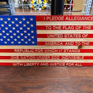 May include: Wooden American flag with the Pledge of Allegiance engraved. The flag features a blue field with gold stars and red and natural wood stripes. The text is in a serif font.