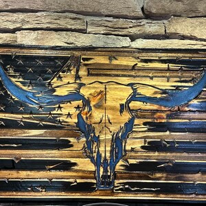 May include: Wooden wall art featuring a carved bull skull superimposed over a distressed American flag design. The flag has blue stars and stripes with a blue bull skull. The wood has a natural, rustic finish.