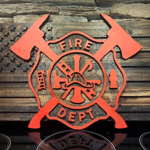 Puede incluir: Arte de pared de madera con un emblema de bombero rojo y hachas cruzadas sobre un diseño de bandera estadounidense desgastada. Las palabras "FIRE DEPT" se muestran de forma destacada. La obra de arte es de madera y tiene un aspecto rústico.