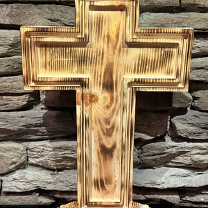Può includere: Una croce di legno con un design a strati e una finitura naturale venata. La croce è montata su una base decorativa. Il legno ha un aspetto bruciato, creando un'estetica rustica. Lo sfondo è un muro di pietra.