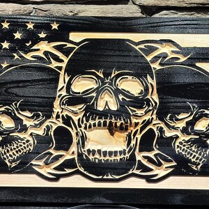 May include: Wooden American flag with carved skulls. The flag features the stars and stripes design, with three skulls in the center. The skulls are intricately carved, adding a unique and edgy aesthetic to the patriotic symbol.