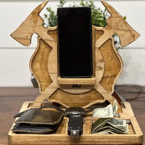 May include: Wooden desk organiser with a phone holder, watch slot, wallet area, and a section for cash. The organiser has a natural wood finish and a unique design with axe-like shapes on the sides. A black smartphone is in the phone holder.