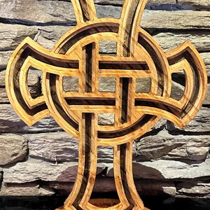 May include: A layered wooden Celtic cross sculpture. The cross features intricate knotwork designs and is constructed from multiple layers of light and dark wood. The sculpture stands on a circular base, showcasing the detailed craftsmanship.