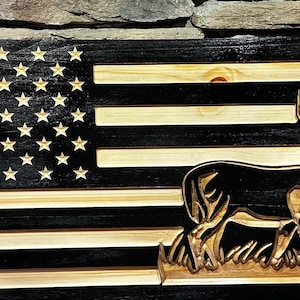 May include: Wooden American flag with a carved deer silhouette. The flag features black and natural wood stripes, with carved stars in the upper left corner. The deer is carved in detail, standing in front of the flag.