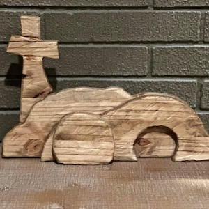 Op de afbeelding: Houten snijwerk dat een kruis en een graf voorstelt. Het kruis staat op een heuvel, met het graf met een open ingang en een grote steen. Het snijwerk is gemaakt van lichtgekleurd hout en heeft een rustieke uitstraling.