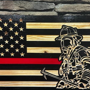 May include: Wooden American flag with a firefighter silhouette. The flag features a black and natural wood design with a red stripe and carved stars. The firefighter is holding a tool.
