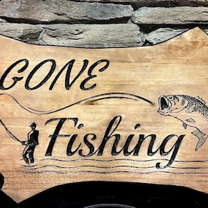 May include: Wooden sign with the words "GONE FISHING" carved in black script. A fisherman silhouette and a leaping fish are also carved into the wood. The sign is a light brown color and has a rustic, natural edge.