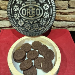 May include: A wooden serving tray in the shape of an Oreo biscuit, with the word "OREO" carved into it. A smaller wooden bowl contains several Oreo biscuits. The biscuits are chocolate with a white cream filling. The tray and bowl are on a red surface.