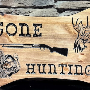 May include: Wooden sign with the words "GONE HUNTING" in black lettering. The sign features a shotgun, a turkey, and a deer head. The sign is made of light brown wood with a dark brown border.