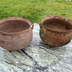 Puede incluir: Dos calderos redondos de hierro fundido resistente con una pátina marrón oxidada y desgastada. Cada recipiente presenta una abertura ancha, un cuerpo redondeado con crestas horizontales, pequeñas asas laterales y tres patas cortas y firmes.