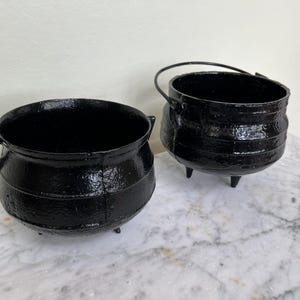 Puede incluir: Dos calderos negros de hierro fundido. Uno tiene un asa, el otro dos asas laterales. Ambos tienen tres patas pequeñas. Los calderos tienen un acabado texturizado y brillante y están sobre una superficie de mármol.