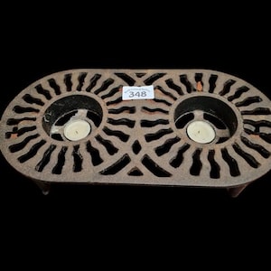 May include: An oval cast iron trivet featuring a decorative sunburst pattern with two circular cutouts, each containing a small white tea light candle. A white rectangular sticker with the number 348 is attached to the center of the metal frame.