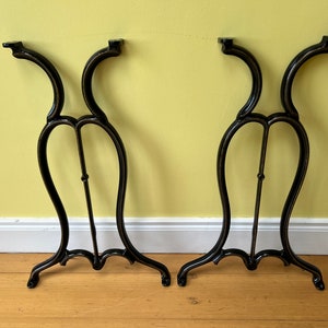 May include: Two black metal table legs with a decorative, curved design. The legs are made of cast iron and have a black finish.