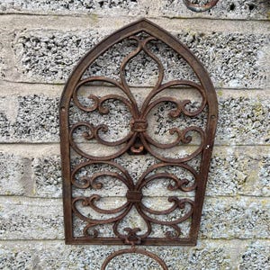 May include: A weathered, ornate, wrought-iron wall-mounted planter holder. The holder features an arched top with scrolling floral designs and a circular ring at the bottom. The metal has a rusty brown patina, mounted on a gray brick wall.
