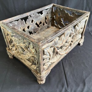 May include: Rectangular metal planter with ornate leaf and floral cutouts. The planter has a weathered, aged appearance with a dark, muted color palette. The sides feature detailed relief carvings, and it rests on small, decorative feet.