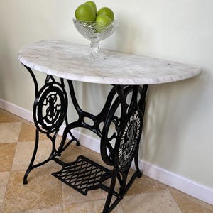 Puede incluir: Una mesa con una tapa de mármol blanco y una base de hierro fundido negro. La base es un soporte de máquina de coser Singer reutilizado con el logotipo de la empresa visible. La mesa tiene aproximadamente 76 cm de altura.