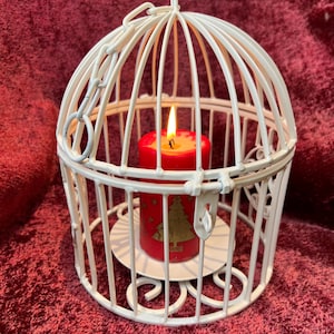 Può includere: Gabbia per uccelli in metallo bianco con una candela rossa all'interno. La candela ha un disegno dorato di un albero di Natale.