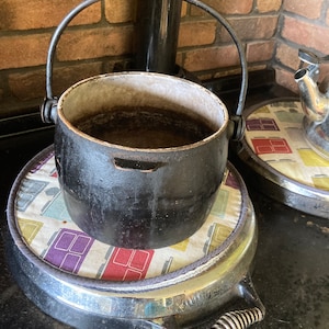 Puede incluir: Una olla de hierro fundido negra con asa se encuentra sobre un trípode redondo plateado con una cubierta de tela estampada. La olla está sobre una superficie negra.