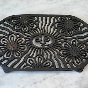 Puede incluir: Salvamanteles de hierro fundido negro con un diseño decorativo de flores y una cara de sol. El salvamanteles tiene forma ovalada y tiene un borde elevado.