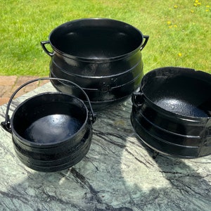 Op de afbeelding: Drie zwarte gietijzeren potten in verschillende maten. De potten hebben een afgeronde vorm met een brede basis en een smalle opening. De grootste pot heeft twee handvatten aan de zijkanten, terwijl de kleinere potten een enkel handvat aan de zijkant hebben. De potten zijn allemaal gemaakt van gietijzer en hebben een gladde, zwarte afwerking.